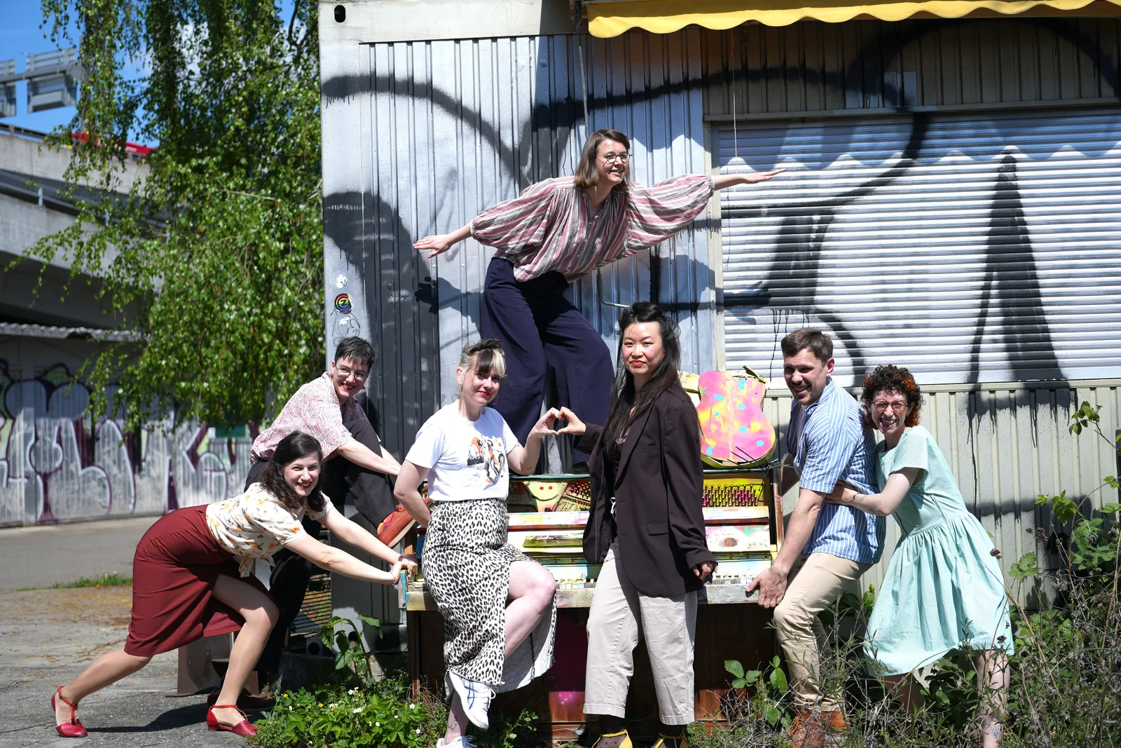 QSXZ team members posing playfully around a colourful piano against a graffiti wall
